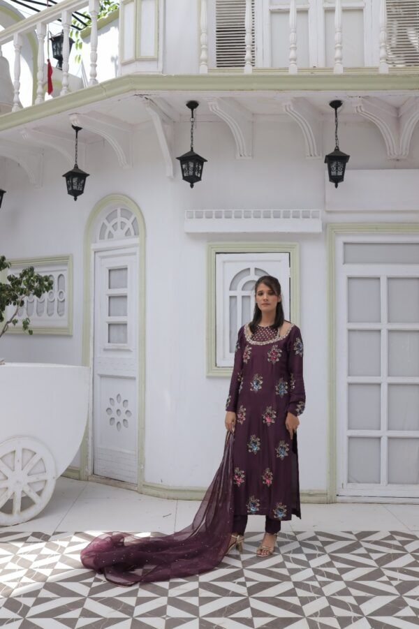 Model in a deep purple pure silk dress with full hand embroidery, stone and sequin embellishments, organza dupatta with four-sided stone lace, and matching pure silk trousers, standing in a white arched backdrop.