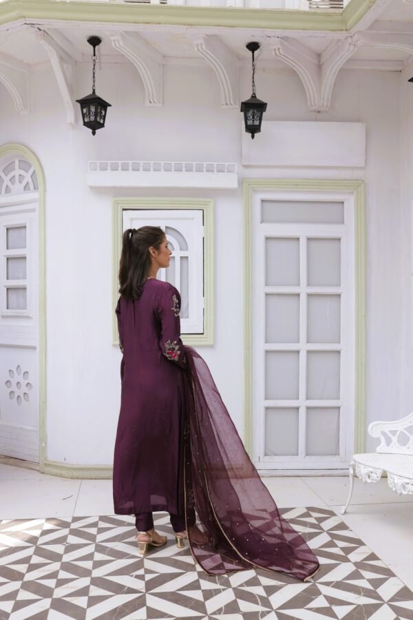 Model in a deep purple pure silk dress with full hand embroidery, stone and sequin embellishments, organza dupatta with four-sided stone lace, and matching pure silk trousers, standing in a white arched backdrop.